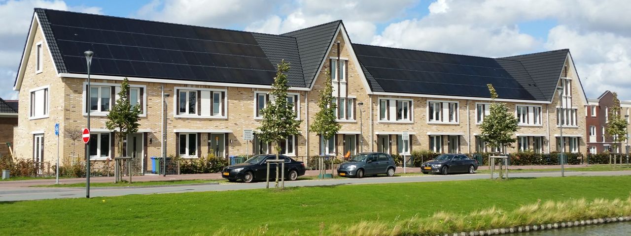 Een rij nieuwe rijtjeshuizen met zonnepanelen op het hele dak, aan een gracht met gras